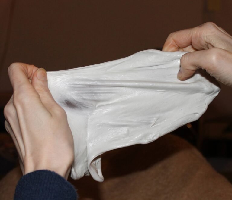 The photo shows two hands extending a gray-white slime into different corners. The extended slime resembles chewing gum, but is much bigger. It is about 25 cm long and 10 cm high. The slime is transparent and opaque at the same time and has a squishy, grippy texture. It makes bubbles that burst with a sharp sound. It is easy to shape because it is not sticky. RC enjoys making slime. For this slime one needs 200g children’s glue, 2g baking soda. then add shaving foam and a few big spoons of contact lens solution (salty solution) A tip: the more contact lens solution, the less sticky the slime will be. Shaving foam makes it fluffy and keeps it that way for a long time.